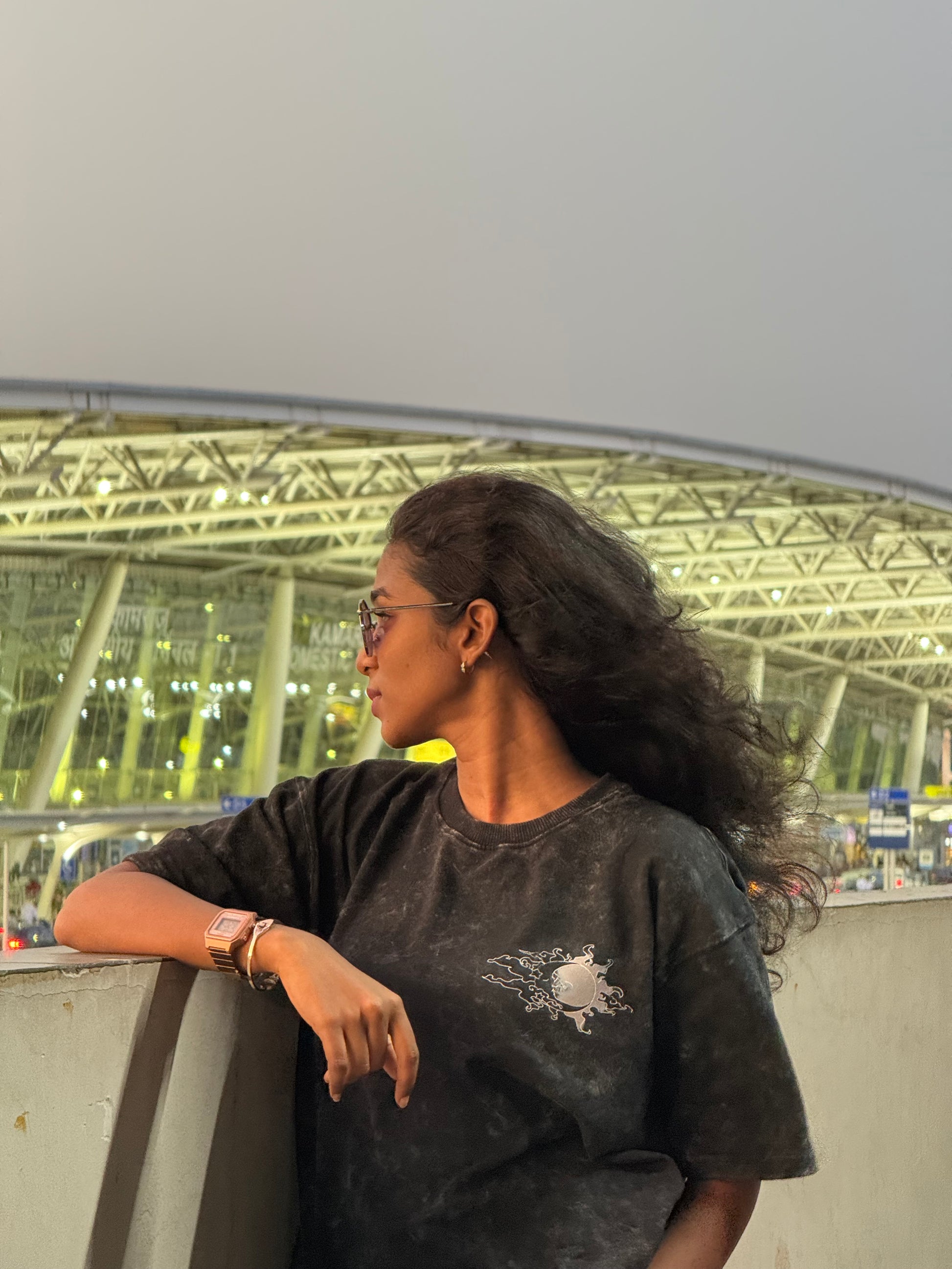 Person wearing a dark shirt with a logo, standing against a blurred indoor background.
