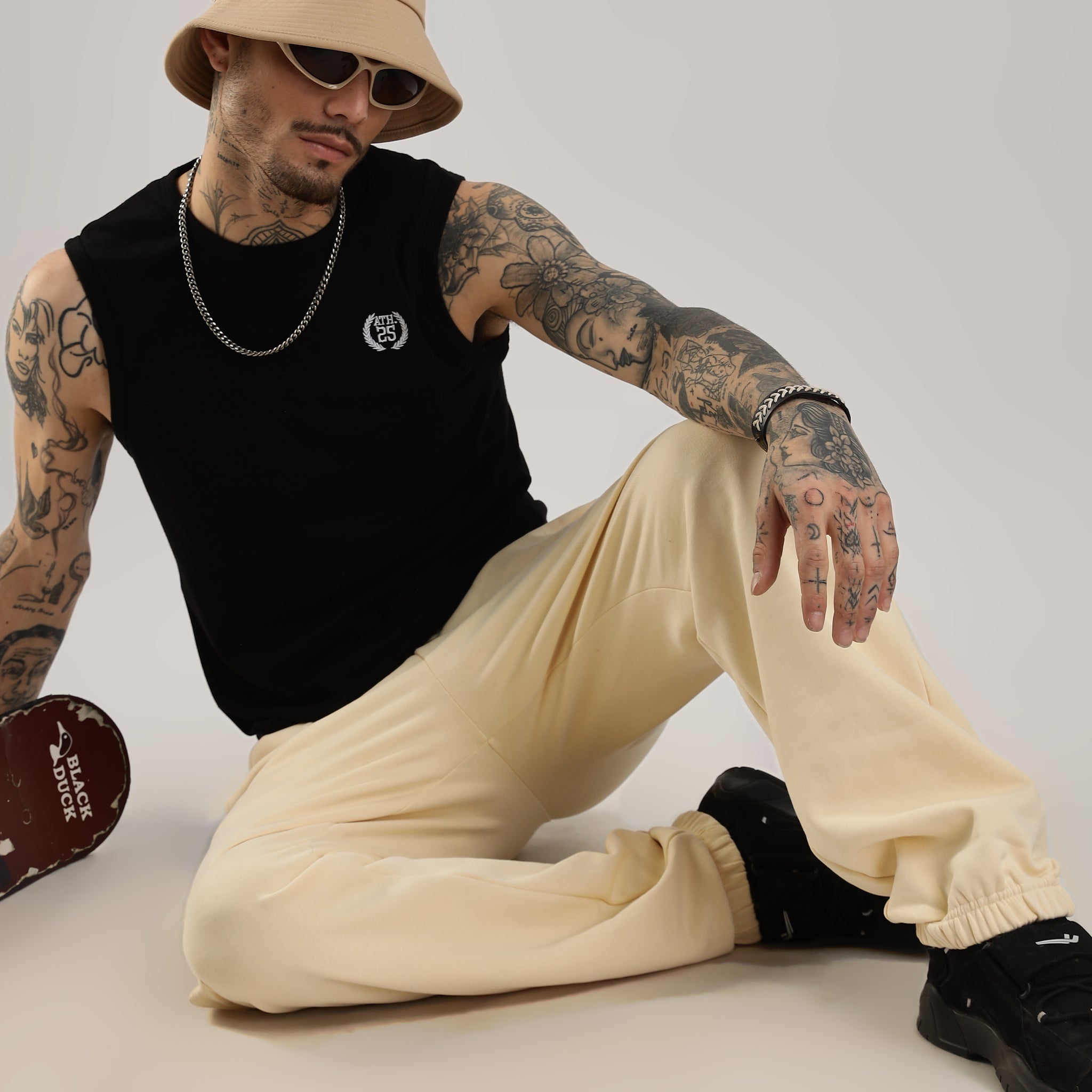 A person wearing beige joggers and a black sleeveless top, accessorized with a hat and sunglasses, sitting against a neutral background.