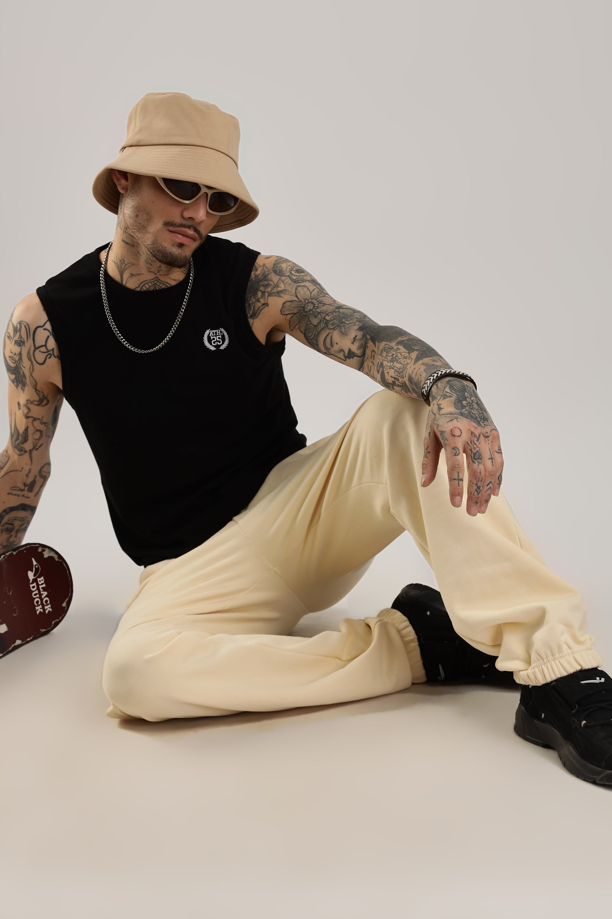 A person wearing beige joggers and a black sleeveless top, accessorized with a hat and sunglasses, sitting against a neutral background.