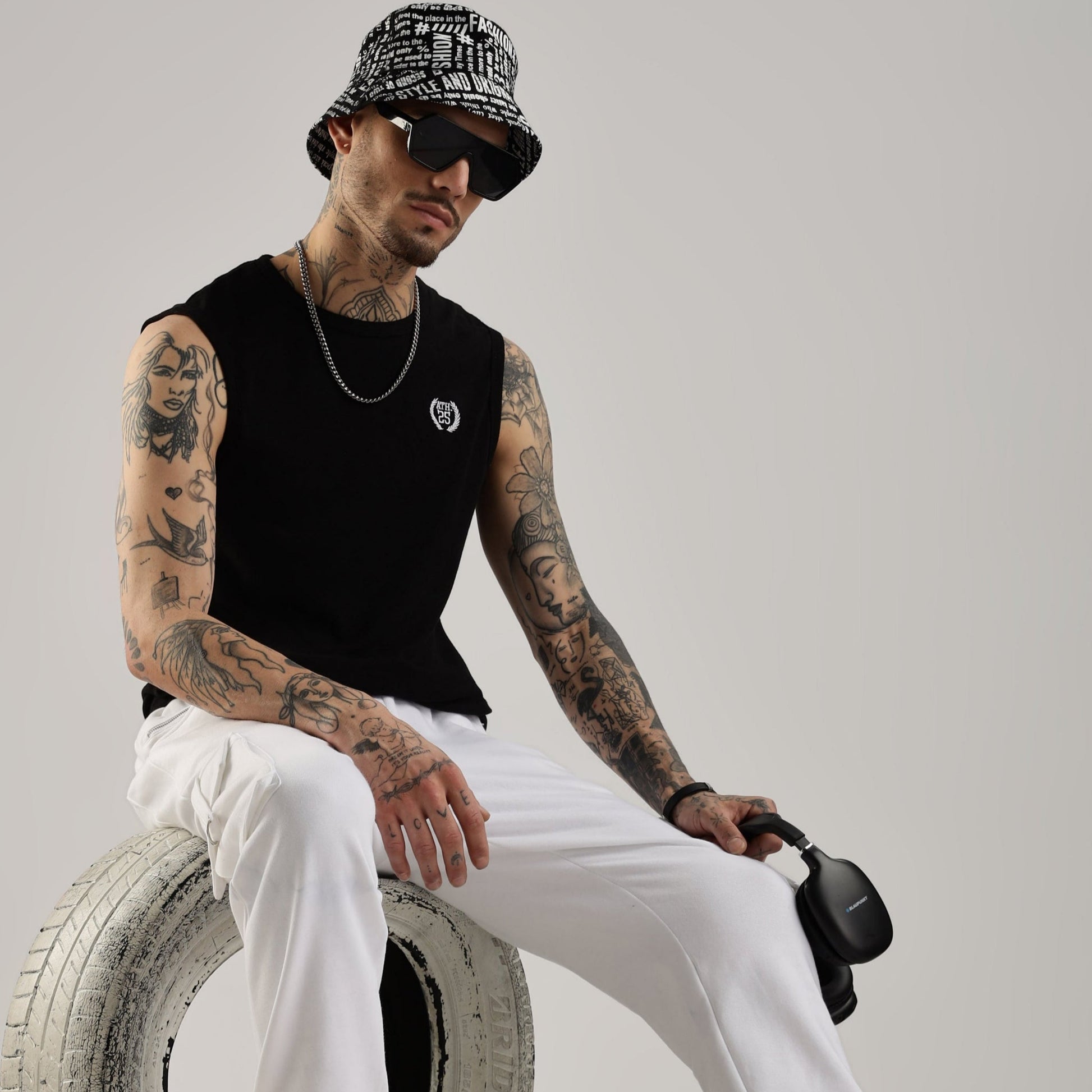 A male model wearing white Versa fit stretch joggers and a black sleeveless top, sitting on a tire with a gray background.