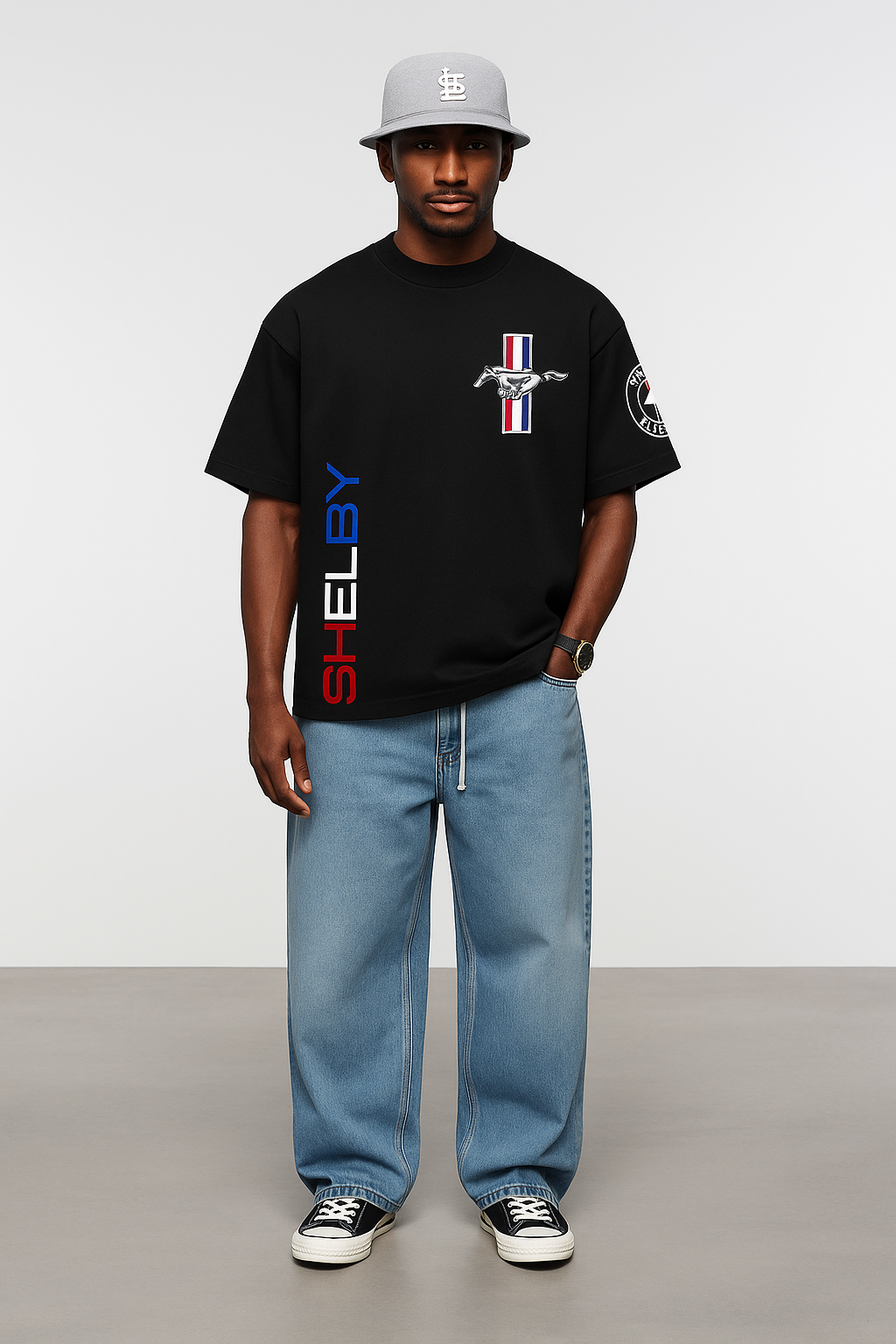 Man wearing a black t-shirt with a logo and text, light blue jeans, and a cap on a plain background