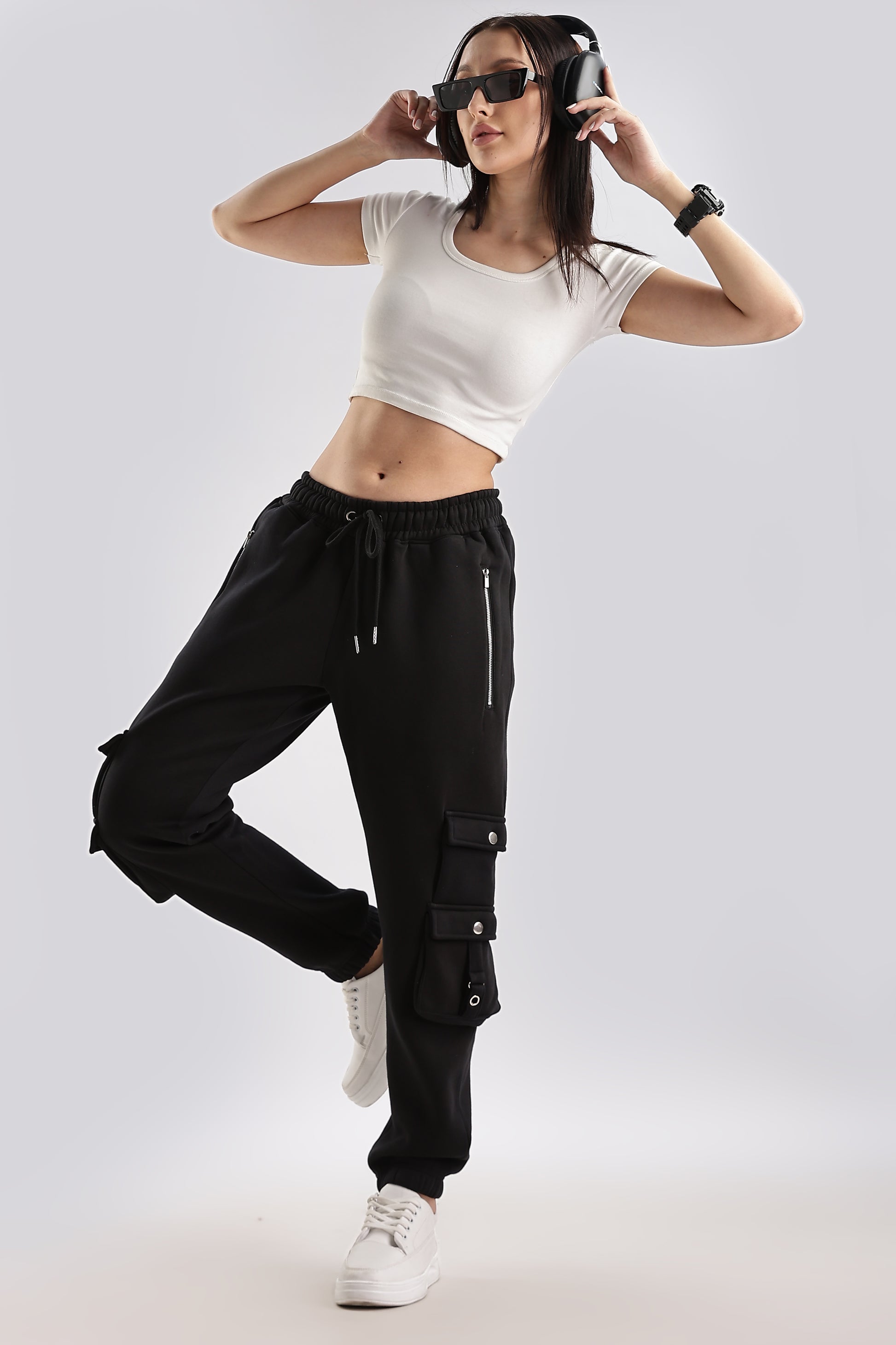 A woman wearing black joggers with adjustable drawstring waistband, ribbed ankle cuffs, and deep side pockets, paired with a white crop top and sunglasses.