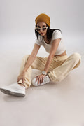 A woman sitting on a white surface wearing beige-core fit stretch joggers with a yellow beanie, white top, and white sneakers.