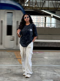 Person wearing a black t-shirt and beige pants standing on a train platform with a blurred train in the background.
