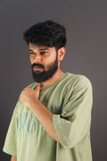 A man wearing an oversized moss green t-shirt with short sleeves and a crew neck, standing against a dark background.