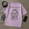 Pink t-shirt with a graphic design and text, placed on a gray surface with headphones and a phone.