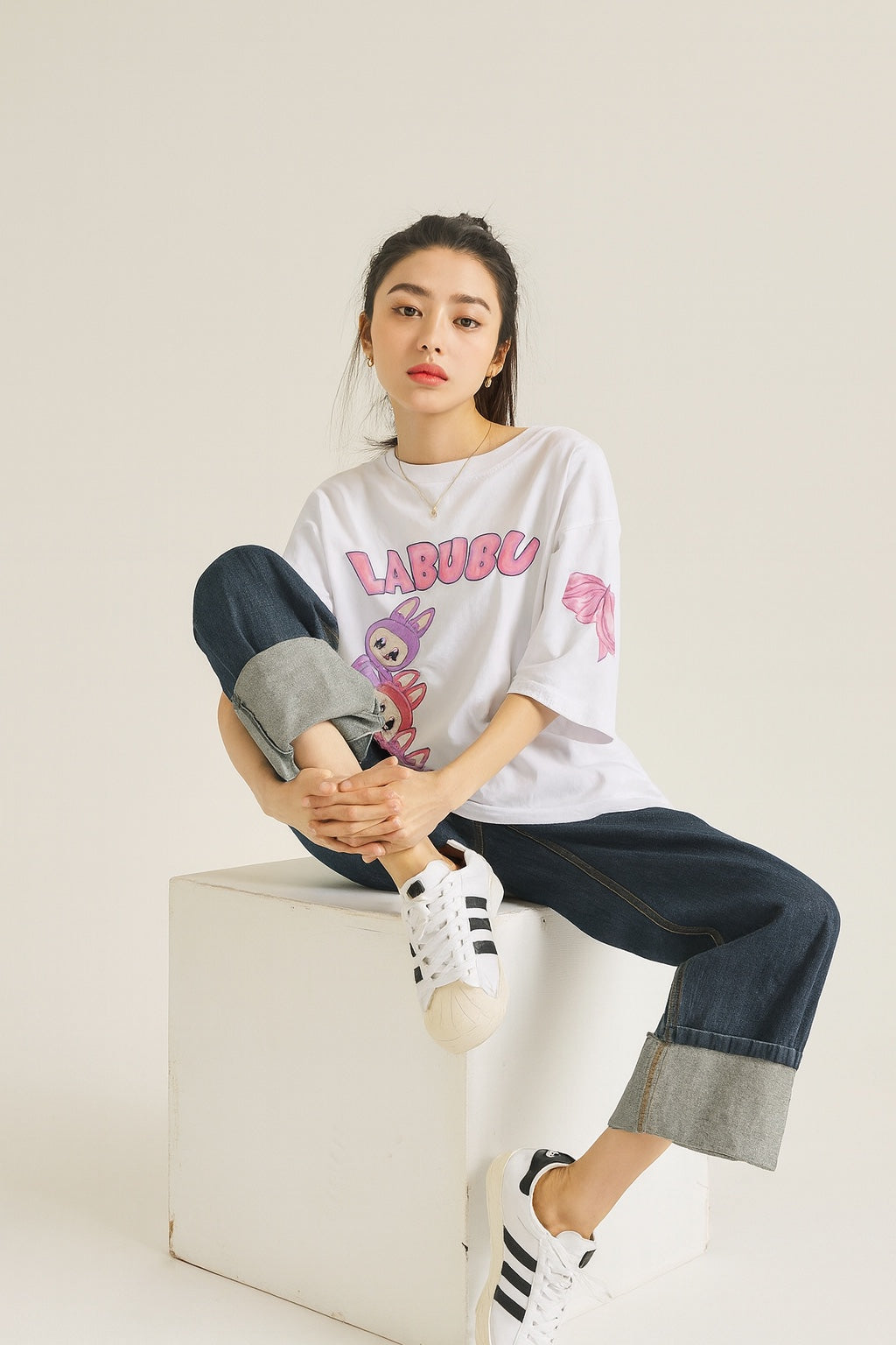 Person wearing a white t-shirt with pink text and graphics, sitting on a white box against a plain background.