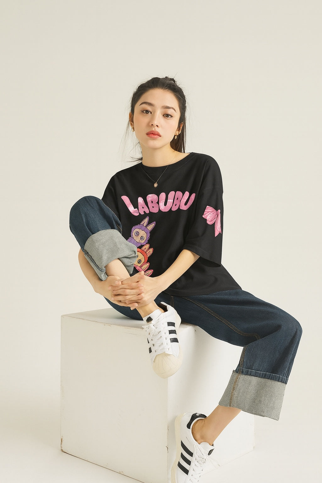Person wearing a black 'LABUBU' t-shirt with colorful graphics, sitting on a white box against a plain background.